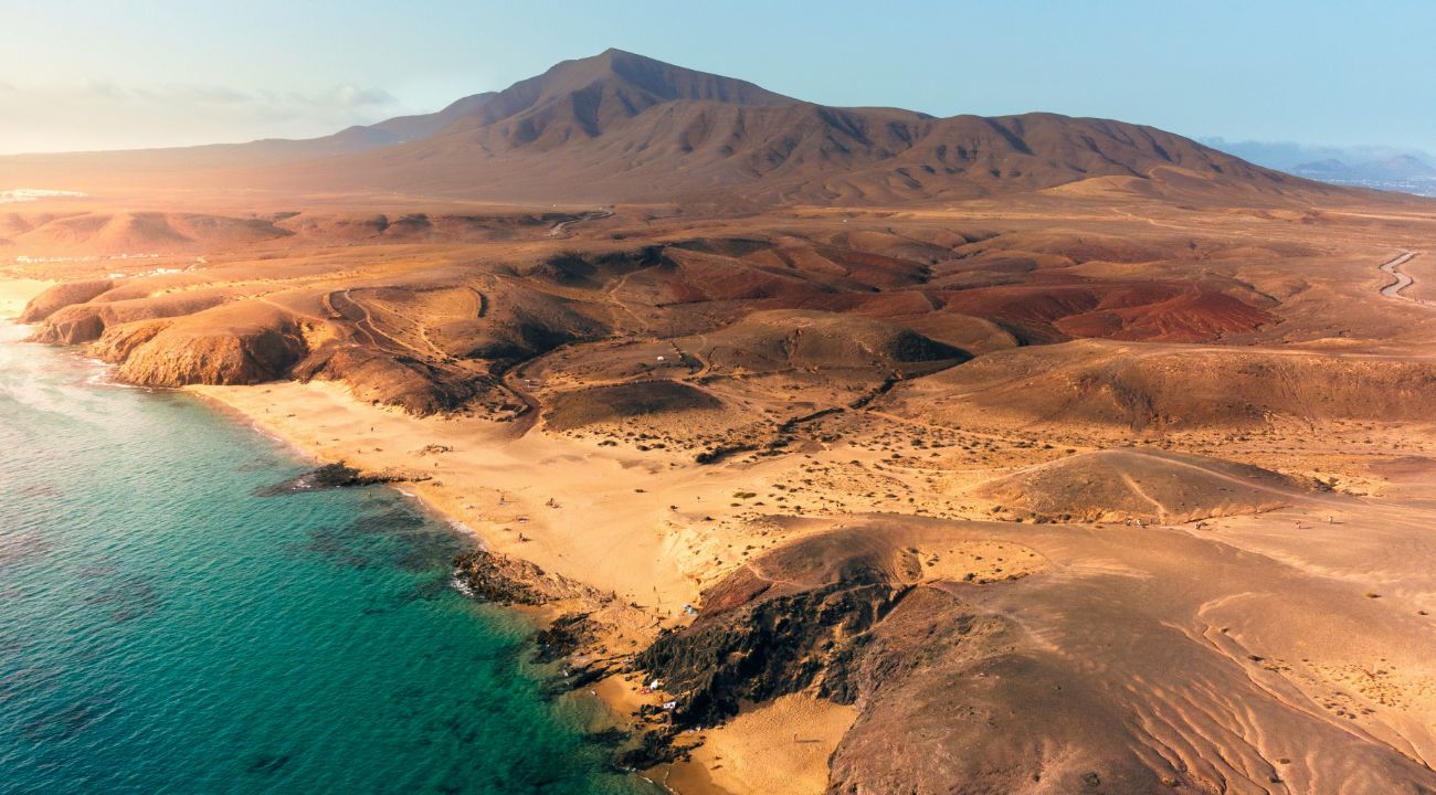 Vista aérea de costa de Lanzarote con playa turquesa, volcanes desérticos y carreteras sinuosas hacia montaña
