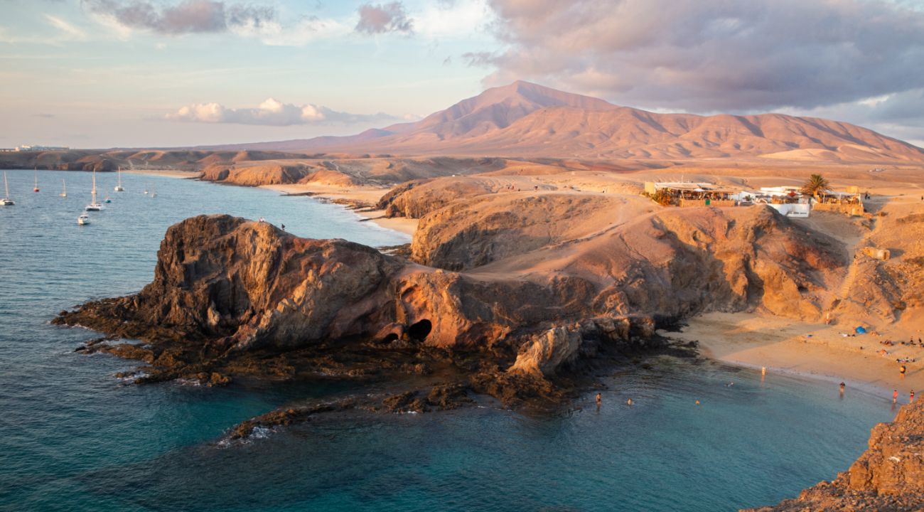 Vista panorámica de la costa volcánica de Lanzarote al atardecer, con formaciones rocosas de basalto, aguas turquesas y un pequeño pueblo blanco al fondo, rodeado de montañas desérticas.